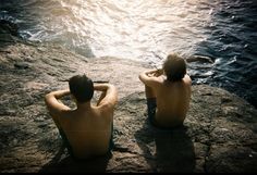 two men sitting on the edge of a cliff looking out at the ocean and water