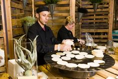 two people preparing food on top of a black plate in front of wooden pallets