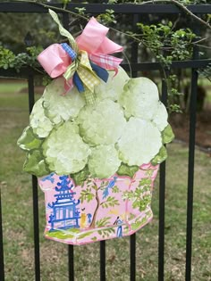 a pink and green cupcake shaped decoration hanging from a black fence with trees in the background