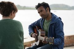 a man sitting on a bench playing an acoustic guitar next to a woman who is looking at him