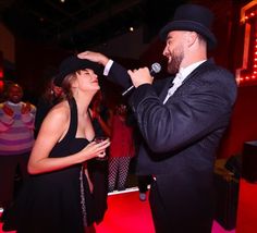 a man in a suit and tie standing next to a woman wearing a hat while holding a microphone