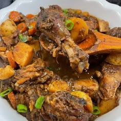 a white bowl filled with meat and vegetables on top of a table next to a wooden spoon