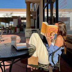 a woman reading a book while sitting at an outdoor table in front of a building