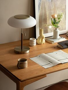 a wooden desk topped with a lamp next to a book and vase filled with flowers