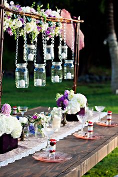 an outdoor table set up for a wedding reception with flowers and hanging mason jars on it