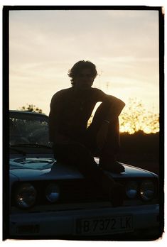 a man sitting on top of a car with the sun setting in the back ground