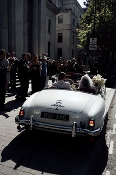 a bride and groom ride in an old car down the street as people look on