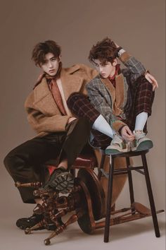 two young men sitting on top of a wooden chair next to each other, one wearing a coat and tie