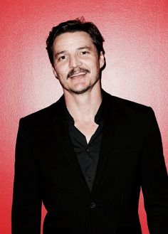 a man with a mustache standing in front of a red wall wearing a black suit