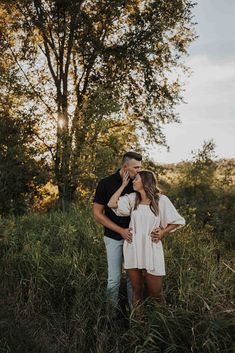 a man and woman standing in tall grass with the sun shining through the trees behind them