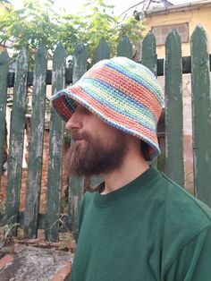 a bearded man wearing a multicolored crocheted hat with a beard standing in front of a fence