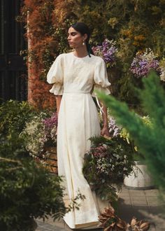 a woman is standing in front of some plants and flowers wearing a white dress with ruffle sleeves