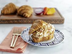 a croissant sandwich on a plate next to a fork and napkin with fruit in the background