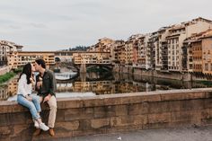 a man and woman sitting on a stone wall next to a river with buildings in the background