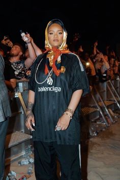 a woman standing in front of a crowd wearing a black shirt and gold head scarf