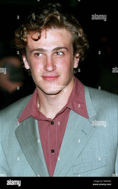 a young man with curly hair wearing a gray suit and red shirt smiling at the camera