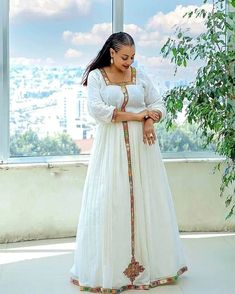 a woman standing in front of a window wearing a white dress with colorful trims