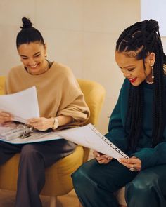 two women sitting on yellow chairs and one is holding a piece of paper while the other looks at something in her hand