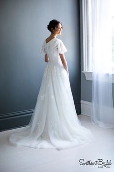 a woman standing in front of a window wearing a white wedding dress with short sleeves