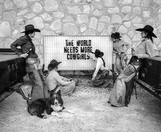 black and white photograph of people with dogs in front of a sign that says the world needs more cowgirls