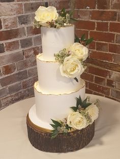 a three tiered white wedding cake with flowers on the top and bottom, sitting in front of a brick wall