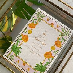 an ornate wedding card with flowers and leaves on it in a gold frame next to a plant