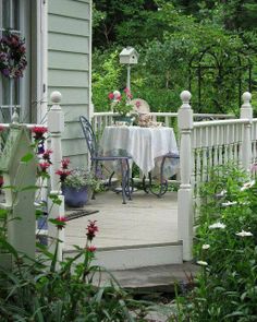 a table and chairs on a porch with flowers in the foreground, and trees in the background