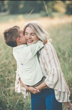 a woman holding a young boy in her arms and kissing him on the cheek while standing in tall grass