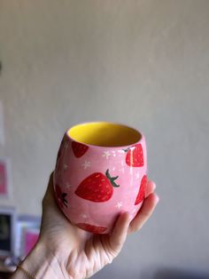 a hand holding a pink and yellow cup with strawberries painted on the outside, in front of a white wall