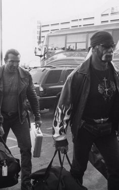 black and white photograph of two men carrying luggage in parking lot with cars behind them