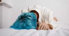 a person with blue hair covering their face on top of a white bed in a room