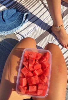 a woman sitting on the beach with a container of watermelon slices in her lap