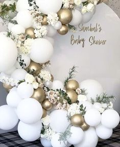 a white and gold balloon arch with flowers on the top, greenery around it