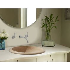 a bathroom sink sitting under a round mirror next to a vase with flowers in it