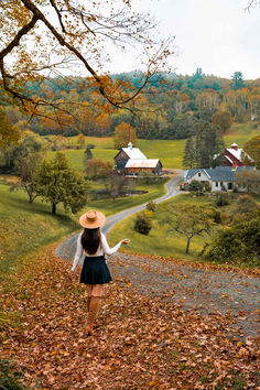 a woman walking down a leaf covered road in the fall with houses and trees behind her