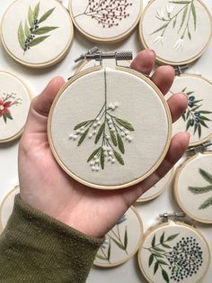 a hand holding a small embroidery kit with flowers and berries on it, in front of several embroidered hoop frames