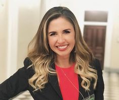 a woman in a red shirt and black blazer smiles at the camera with her hands on her hips