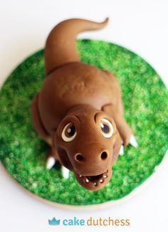 a chocolate dinosaur cake sitting on top of a green grass covered plate with the face of a dog