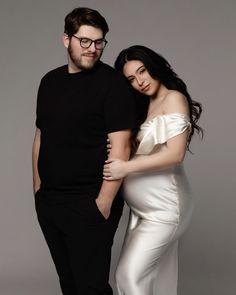 a man standing next to a woman in a white dress on a gray background wearing glasses