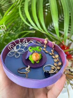 a hand holding a purple tray with various jewelry on it and plants in the background