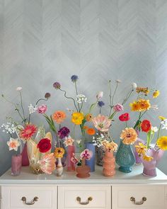 an assortment of colorful flowers in vases on top of a white dresser against a chevron wall