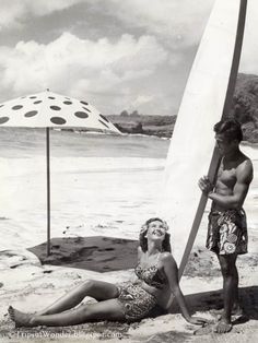 a man and woman sitting on the beach with a surfboard in front of them