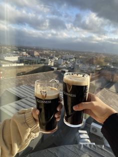 two people toasting with dark beer on top of a building overlooking a cityscape