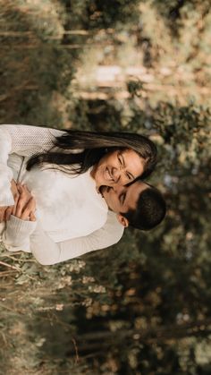 a man and woman standing next to each other in front of some trees with their arms around each other