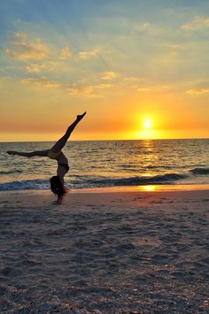 a woman doing a handstand on the beach at sunset with her feet in the air