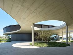 the circular building has two trees in front of it and is surrounded by concrete pillars