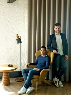 two men sitting on chairs in front of a curtained window with a lamp next to them