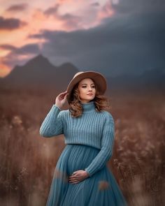 a woman in a blue dress and brown hat poses for a photo while standing in tall grass