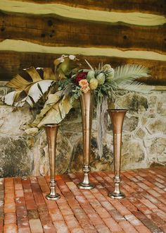 two gold vases with flowers are sitting on a brick floor next to a stone wall