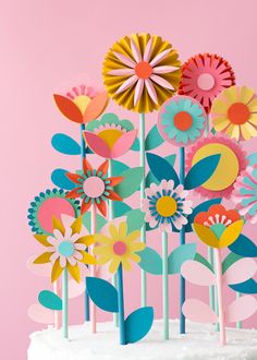 colorful paper flowers on top of a cake with white frosting and pink icing